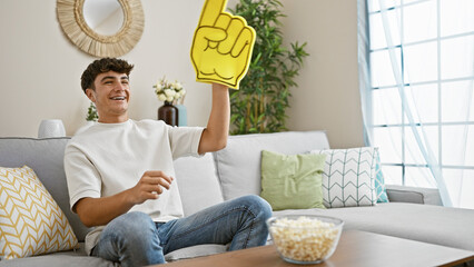 Excited young hispanic male teenager, avid soccer team fan, joyfully watching the match on tv,...