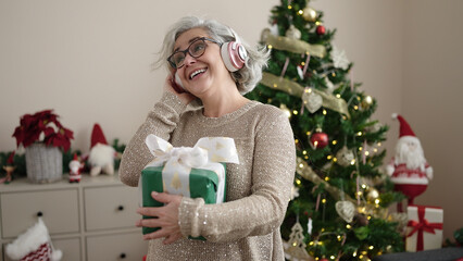 Obraz na płótnie Canvas Middle age woman with grey hair listening to music standing by christmas tree at home