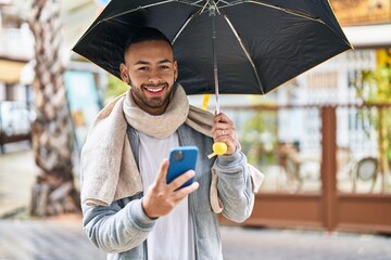 African american man using smartphone holding umbrella at street