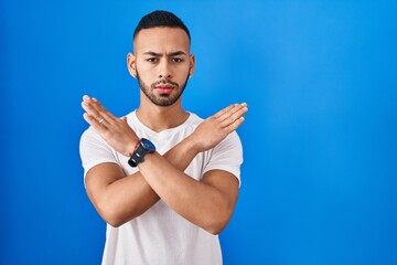 Young hispanic man standing over blue background rejection expression crossing arms doing negative...