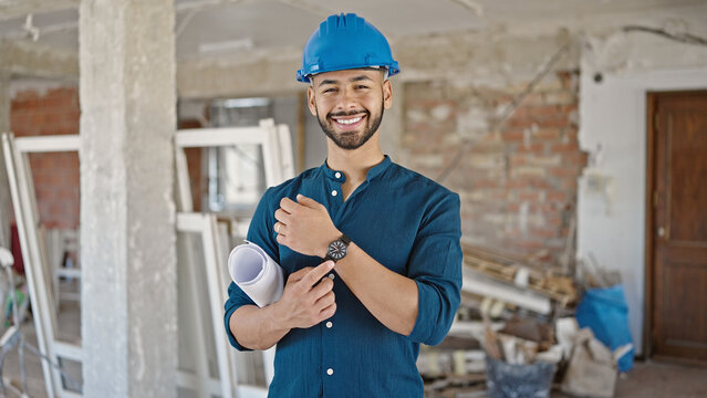 Young hispanic man architect wearing hardhat holding blueprints pointing at wrist watch at construction site