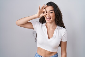 Young teenager girl standing over white background doing ok gesture with hand smiling, eye looking through fingers with happy face.