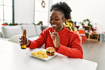 African american woman eating hamburger drinking beer sitting on table by christmas tree at home