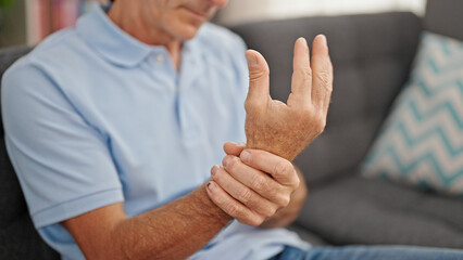 Middle age man sitting on sofa suffering for wrist pain at home - obrazy, fototapety, plakaty