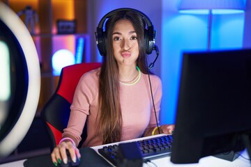Young hispanic woman playing video games puffing cheeks with funny face. mouth inflated with air, crazy expression. - obrazy, fototapety, plakaty