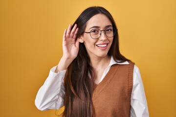 Young brunette woman standing over yellow background wearing glasses smiling with hand over ear listening an hearing to rumor or gossip. deafness concept.