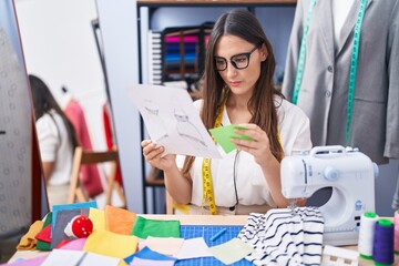 Young beautiful hispanic woman tailor looking clothing design holding cloth at clothing factory