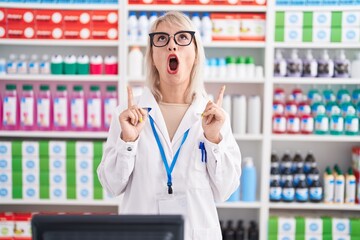 Young caucasian woman working at pharmacy drugstore amazed and surprised looking up and pointing with fingers and raised arms.