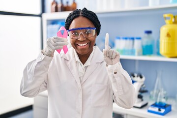 Naklejka premium African american woman working at scientist laboratory holding pink ribbon surprised with an idea or question pointing finger with happy face, number one
