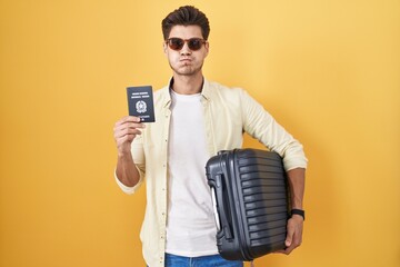 Young hispanic man holding suitcase going on summer vacation holding italian passport puffing cheeks with funny face. mouth inflated with air, catching air. - obrazy, fototapety, plakaty
