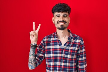 Fototapeta premium Young hispanic man with beard standing over red background showing and pointing up with fingers number two while smiling confident and happy.
