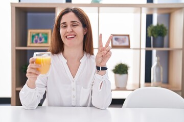 Brunette woman drinking glass of orange juice smiling with happy face winking at the camera doing victory sign with fingers. number two.