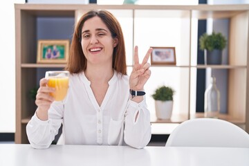 Brunette woman drinking glass of orange juice smiling with happy face winking at the camera doing...