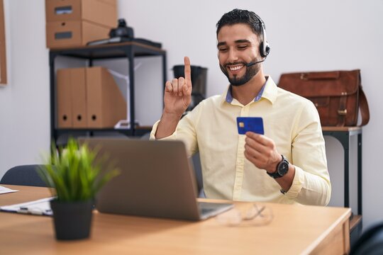 Hispanic young man wearing call center agent headset doing online shopping smiling with an idea or question pointing finger with happy face, number one