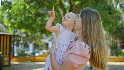 Confident mother holding her cheerful daughter in arms, standing at park looking up, enjoying...