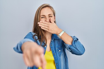 Young blonde woman standing over blue background laughing at you, pointing finger to the camera with hand over mouth, shame expression