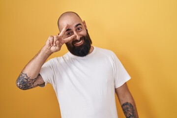 Young hispanic man with beard and tattoos standing over yellow background doing peace symbol with fingers over face, smiling cheerful showing victory