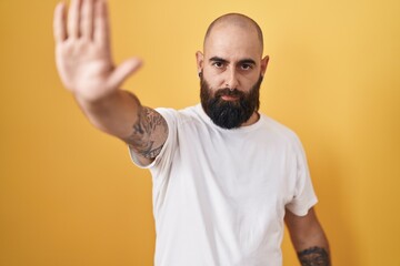 Young hispanic man with beard and tattoos standing over yellow background doing stop sing with palm...