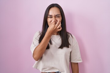 Young hispanic woman standing over pink background smelling something stinky and disgusting,...