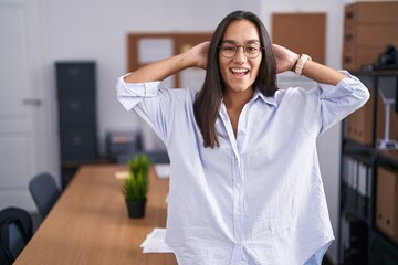 Young hispanic woman at the office relaxing and stretching, arms and hands behind head and neck smiling happy