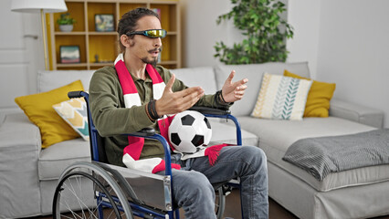 Young hispanic man watching soccer game using virtual reality glasses sitting on wheelchair at home