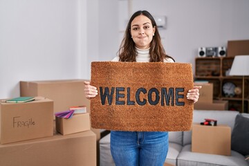 Young hispanic woman holding welcome doormat at new home puffing cheeks with funny face. mouth inflated with air, catching air. - obrazy, fototapety, plakaty