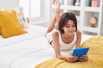Adorable hispanic girl using touchpad sitting on bed at bedroom