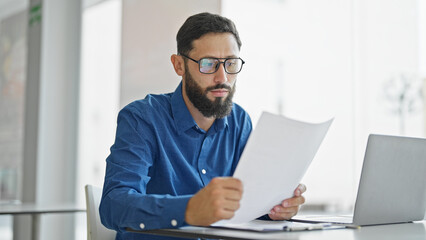 Young hispanic man business worker wearing glasses reading document at the office