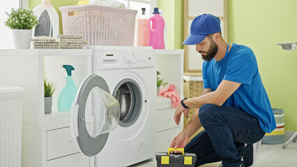 Young hispanic man technician repairing washing machine at laundry room - obrazy, fototapety, plakaty