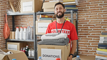 Young hispanic man volunteer holding donation package with clothes smiling at charity center
