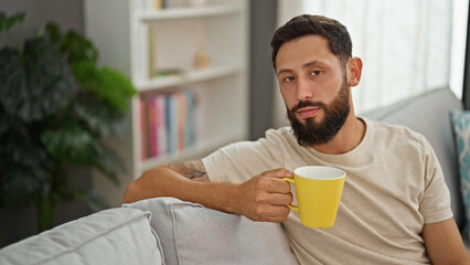 Young hispanic man drinking coffee sitting on sofa at home