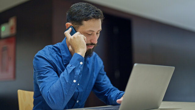 Young hispanic man business worker using laptop talking on smartphone at the office