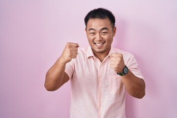 Chinese young man standing over pink background very happy and excited doing winner gesture with arms raised, smiling and screaming for success. celebration concept.