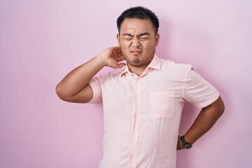 Chinese young man standing over pink background suffering of neck ache injury, touching neck with hand, muscular pain