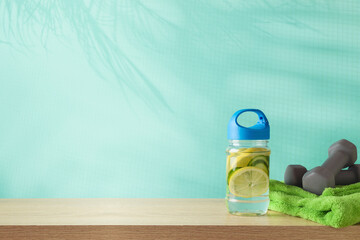 Fitness background with infused water bottle, towel and dumbbells on wooden table over blue background with palm tree shadows - 672792827