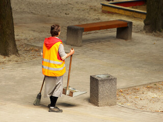 A woman janitor in orange overalls with a broom and dustpan stands on the street near the garbage...
