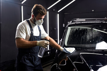 young man cleaning car with cloth and detergent liquids, car detailing or valeting concept. In auto repair shop, side view on male in protective mask, gloves and uniform