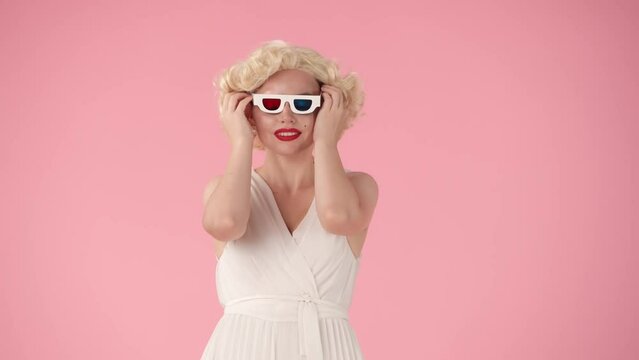 Woman puts on 3d glasses, takes popcorn and watches a movie. Woman eating popcorn, wondering about the plot of the movie. Woman in the look of in a studio on a pink background.