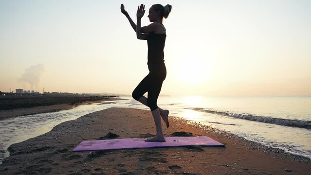 Slender woman doing yoga exercises on  seashore near the waves, dawn time, sun over  horizon