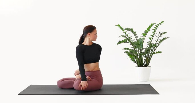 A woman leading a healthy lifestyle and practicing yoga, performs the Parivritta Padmasana exercise, twisting in the lotus position, trains alone in sportswear in a room sitting on a mat