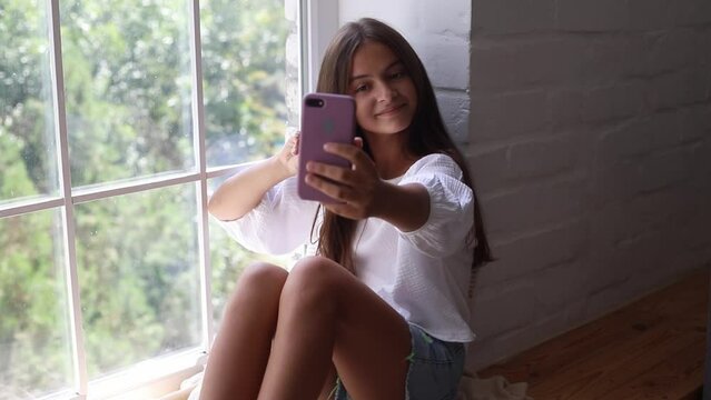 Happy pretty teenage brunette girl making selfie by smartphone sitting at windowsill