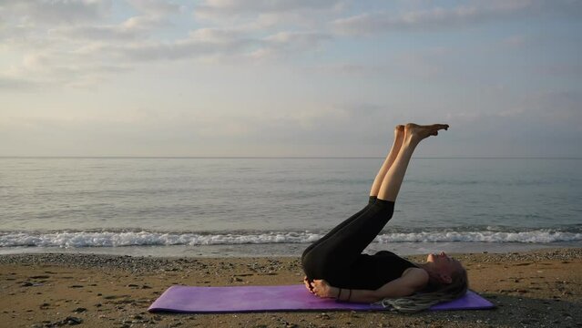 Slender woman doing yoga exercises on  seashore near the waves, dawn time, sun over  horizon