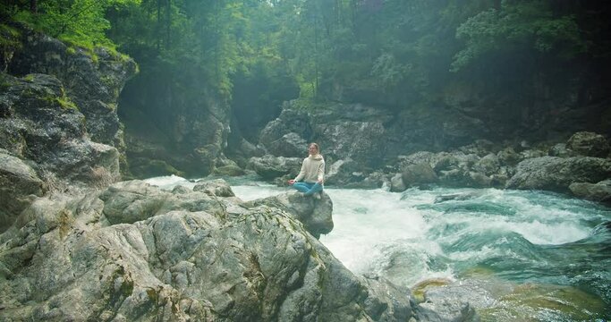 Young woman meditating by the river in early foggy morning. Girl resting in the wild landscape with fast flowing mountain stream.