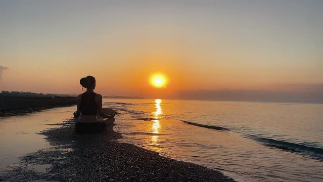 Silhouette of slender woman sitting on the seashore among the waves at dawn, meditating in the lotus position