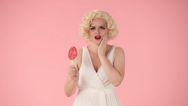 A woman holds her cheek while experiencing a toothache. Unhappy woman in the image of with a lollipop on a stick in her hand in the studio on a pink background.