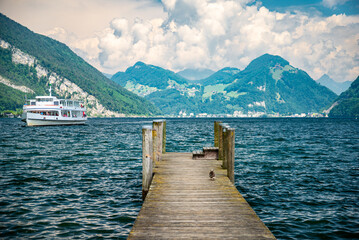 A ship cruises on Lake Lucerne with rippled waters, featuring a pier in the foreground. Enjoy the cloudy skies, deep blue waters, and lush green Alpine hills. Scenic Swiss Mountain Landscape.