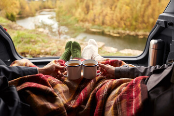 travel, tourism and camping concept - view to river from car trunk with couple under blanket toasting tea cups - obrazy, fototapety, plakaty