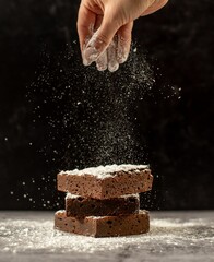 Hand sprinkled sugar onto three stacked brownies covered in powdered sugar - obrazy, fototapety, plakaty