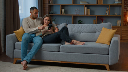 Diverse man and woman interracial couple homeowners relax on sofa talk video call communicate...