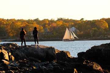 Two people standing on the rocky shore of a lake admiring the view of sailboats in the distance.